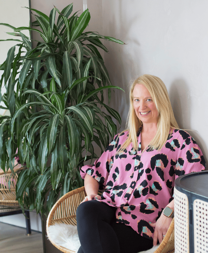 Portrait of Claire Elmes on modern chair with leafy green plant in background.
